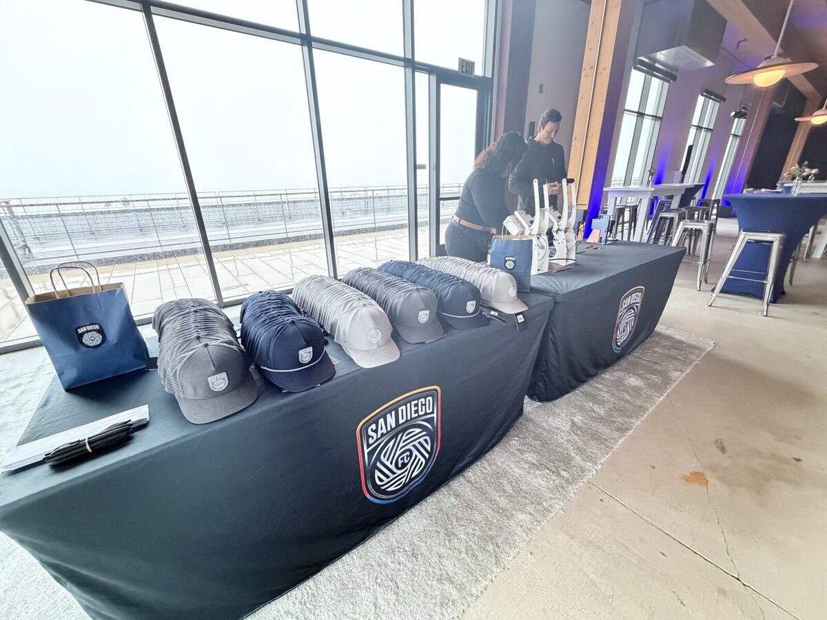 Custom-printed caps laid out on a draped table beside floor-to-ceiling windows overlooking San Diego Bay